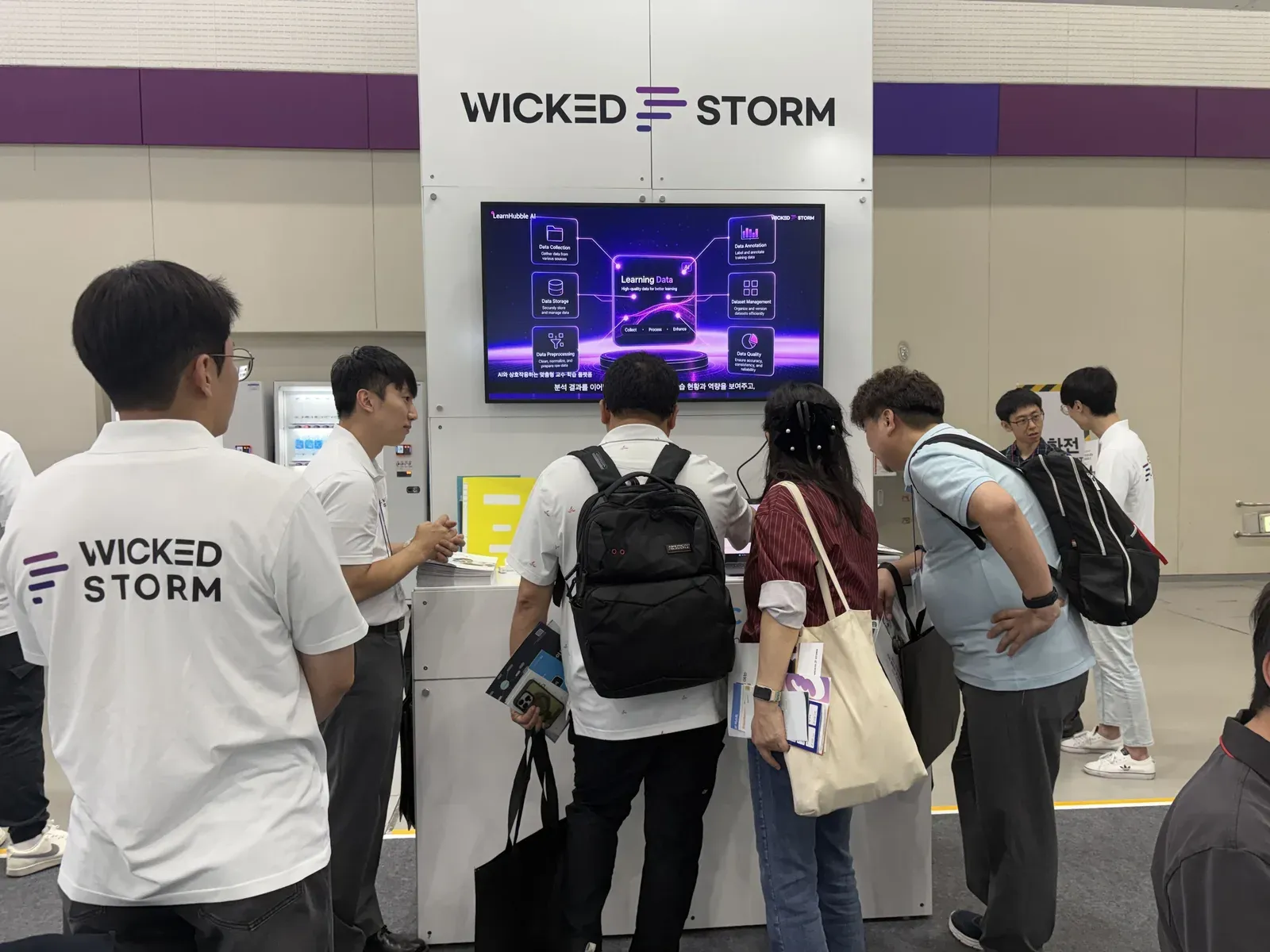 LearnHubble AIの紹介画面の前で、Wicked Stormのスタッフが来場者に説明する様子