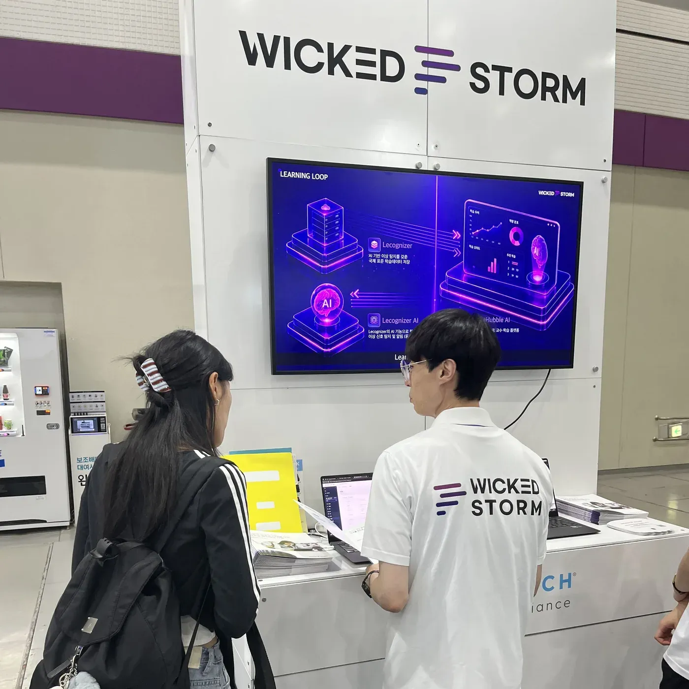 Learning Loop(学びの好循環)の映像が流れる画面の前で、Wicked Stormのスタッフが来場者に説明する様子