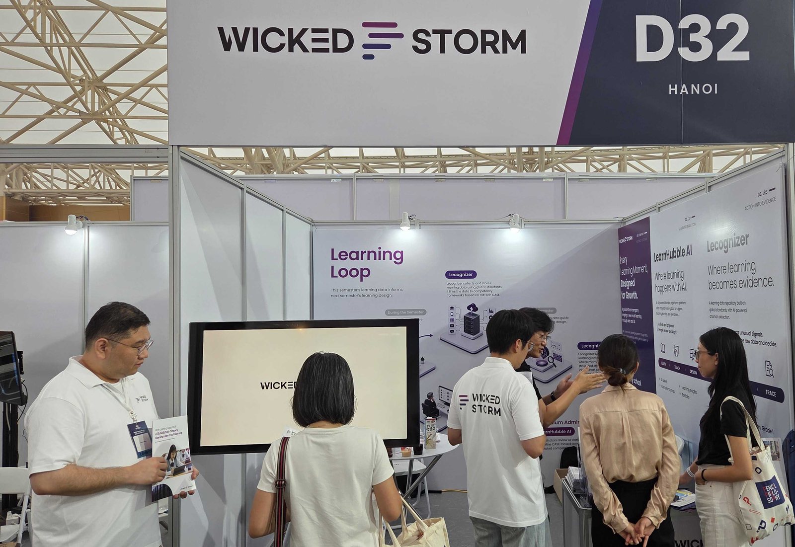 ‘VIETEDU 2026’ 위키드스톰 D32 부스에서 WICKED STORM 티셔츠를 입은 직원이 벽면을 가리키며 관람객들에게 제품을 설명하고, 왼쪽에서는 다른 직원이 소개 자료를 들고 관람객을 맞는 모습